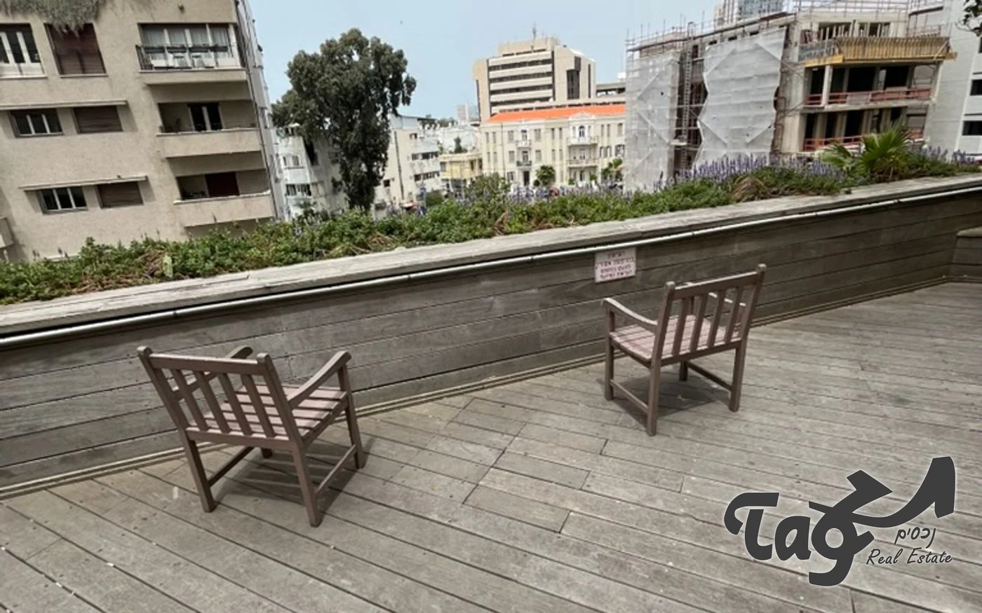 מרפסת עץ עם כיסאות גן וערוגה, ומבט לבניינים בסביבה