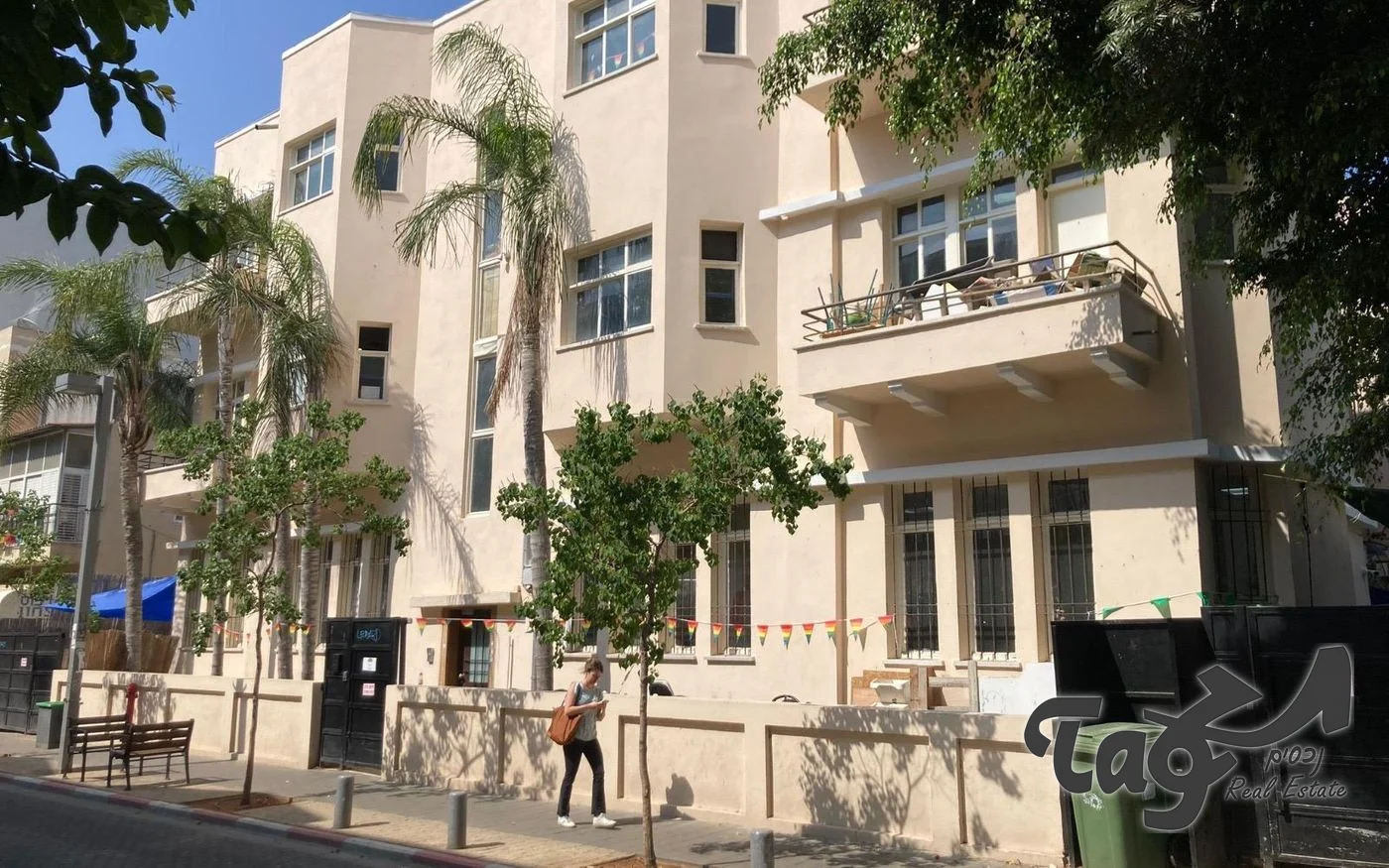 חזית הבניין לשימור במזא״ה במבט מהרחוב, עם מרפסת בעלת מעקה מתכת מימין ועצי דקל משמאל