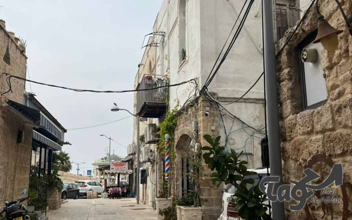 מדרחוב צר ביפו עם בתי אבן ובניינים מטויחים משני הצדדים, מרפסת ברזל וצמחייה על הקירות