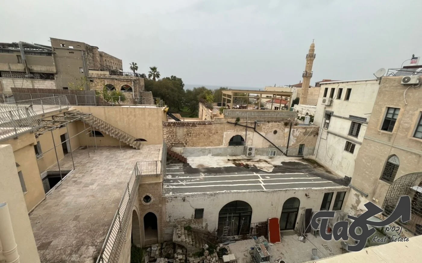 מבט מהגג אל החצר הפנימית והגגות הסמוכים: גג שטוח עם מדרגות חיצוניות וקשתות בקומת הקרקע, ובמרחק צריח של מסגד