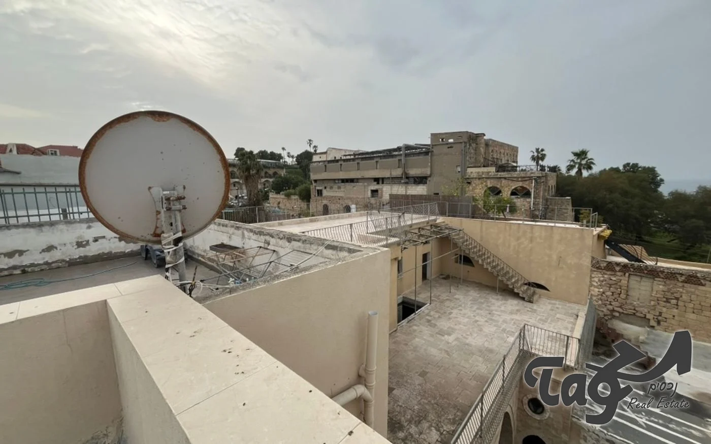 גג שטוח עם מדרגות חיצוניות ומעקה, ומאחוריו גגות יפו ועצי דקל