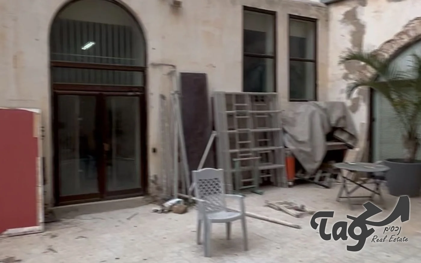 חצר פנימית עם קיר מטויח, קשת גבוהה ודלת זכוכית במסגרת כהה, וחומרי בנייה וכיסא על הריצוף