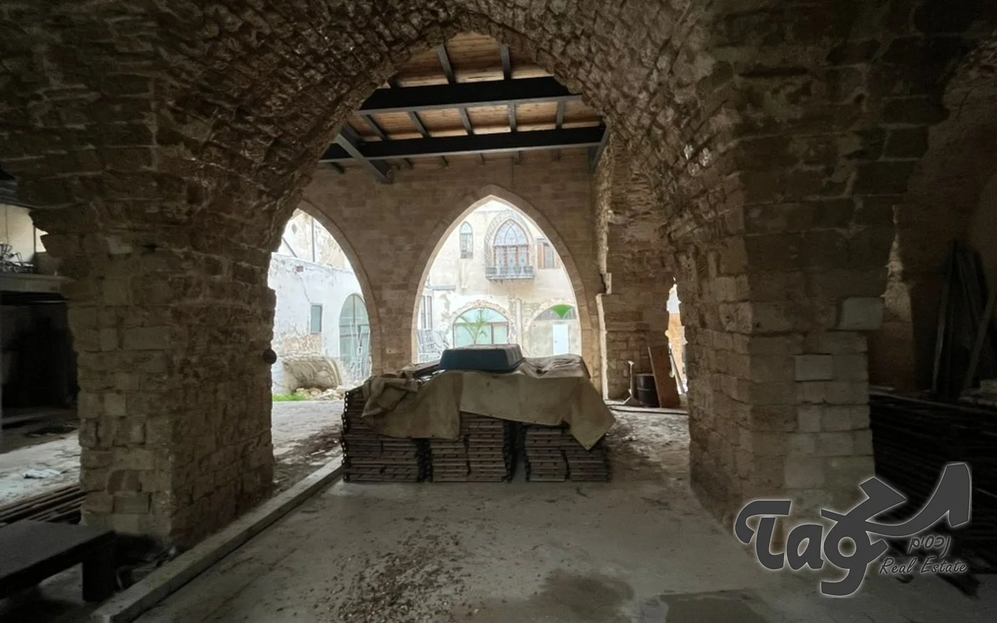 אולם קמרונות אבן בקומת הקרקע: קשתות עגולות מאבן וחלונות בקשת מחודדת הפונים לחצר, עם חומרי בנייה על הרצפה